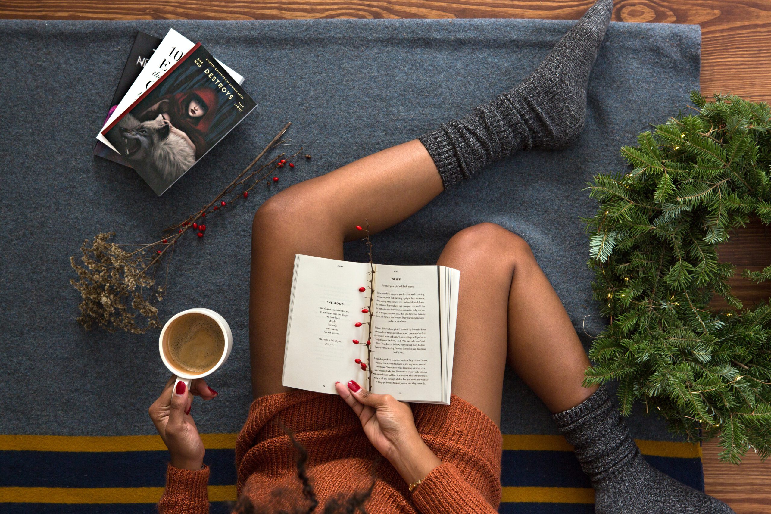 Eine Frau hält in der rechten Hand ein Buch und in der linken Hand eine Tasse Kaffee. Sie trägt dicke Socken, neben ihr liegen Bücher.